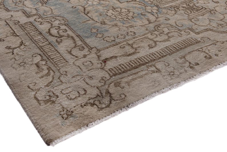May include: Close-up of the edge of a light brown rug with a faded blue and brown floral pattern. The rug has a fringe border.