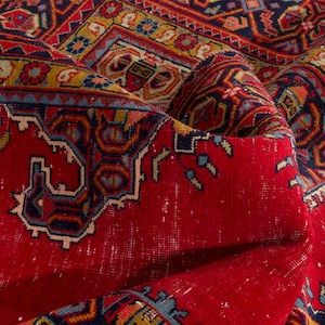 May include: Close-up of a vibrant, folded red rug with intricate patterns. The rug features a central red field with detailed borders in blue, yellow, and other colors. The design includes geometric shapes and floral motifs, showcasing a traditional style.