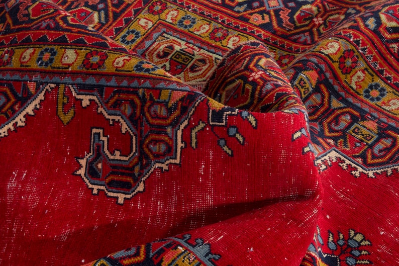 May include: Close-up of a vibrant, folded red rug with intricate patterns. The rug features a central red field with detailed borders in blue, yellow, and other colors. The design includes geometric shapes and floral motifs, showcasing a traditional style.