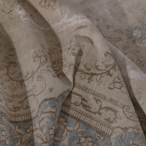 May include: A close-up of a beige and blue patterned rug with a floral design. The rug is folded in half, showing the intricate details of the pattern.