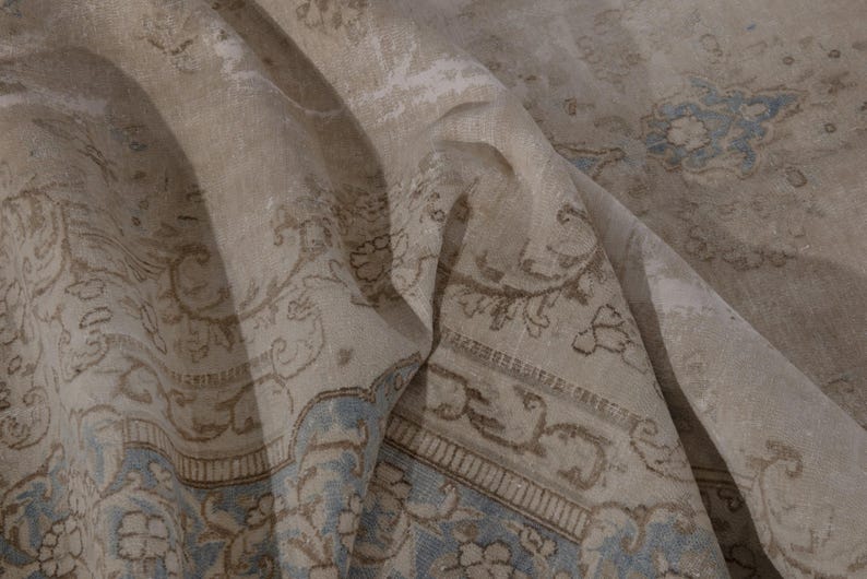 May include: A close-up of a beige and blue patterned rug with a floral design. The rug is folded in half, showing the intricate details of the pattern.