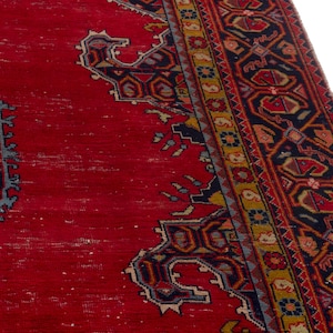 May include: A section of a red oriental rug with intricate geometric patterns in navy blue, gold, and other colors. The rug has a distressed appearance, with visible wear and tear, and a detailed border.