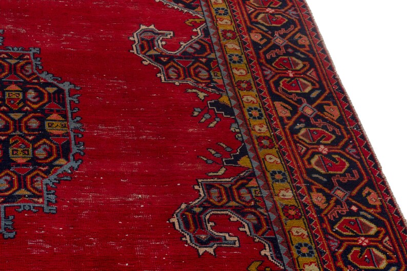 May include: A section of a red oriental rug with intricate geometric patterns in navy blue, gold, and other colors. The rug has a distressed appearance, with visible wear and tear, and a detailed border.