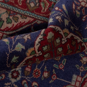 Peut inclure: Gros plan sur un tapis roulé à motifs complexes. Le tapis présente un fond bleu profond avec des accents rouges et beiges. Des motifs floraux et géométriques détaillés sont visibles, créant une apparence riche et texturée. Le design du tapis suggère un style oriental traditionnel.