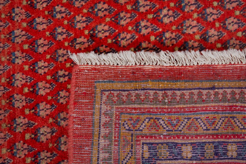 Puede incluir: Primer plano de una alfombra oriental roja con un patr&oacute;n detallado. La alfombra presenta un dise&ntilde;o floral repetido en tonos azules y beige. El borde tiene patrones geom&eacute;tricos y flecos.
