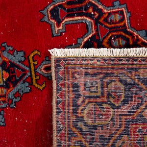 May include: Close-up of a red rug with a detailed geometric pattern in navy blue, teal, and gold. The rug has a fringed edge and a traditional design, showcasing intricate motifs and a rich color palette.