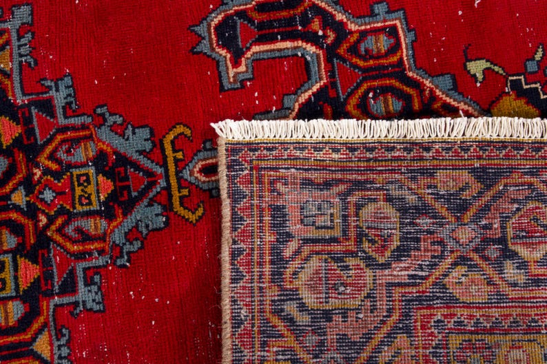 May include: Close-up of a red rug with a detailed geometric pattern in navy blue, teal, and gold. The rug has a fringed edge and a traditional design, showcasing intricate motifs and a rich color palette.