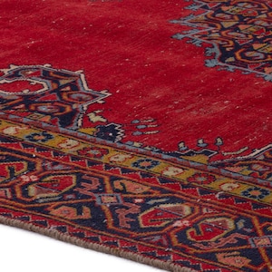 May include: A section of a red oriental rug with intricate geometric patterns in navy blue, gold, and other colors. The rug has a distressed appearance, with visible wear and tear, adding to its vintage charm.