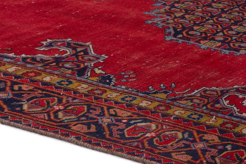 May include: A section of a red oriental rug with intricate geometric patterns in navy blue, gold, and other colors. The rug has a distressed appearance, with visible wear and tear, adding to its vintage charm.