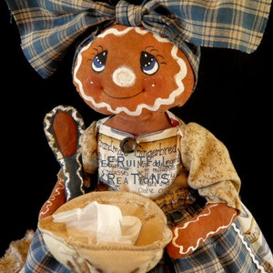 Könnte beinhalten: Eine Lebkuchenpuppe in einem blau-braunen karierten Kleid und einem passenden Stirnband. Die Puppe hält eine Schüssel und hat ein bemaltes Gesicht mit blauen Augen und einem Lächeln. Das Kleid der Puppe trägt den Text "Grandma's Gingerbread".