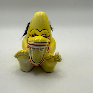 May include: A yellow ceramic figurine of a cartoon-like creature with black hair, red accents, and a wide open mouth. The figurine has a smiling expression and is sitting down.