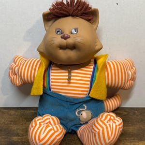 May include: A vintage plush cat doll with brown fur, sparkling eyes, and a tuft of brown hair. It wears a striped orange and white shirt, blue overalls, and a yellow vest. The doll has a small mouse applique on the overalls.