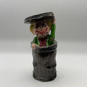 May include: A vintage ceramic figurine of a clown popping out of a trash can. The clown has a wide red smile, blonde hair, and is wearing a green jacket over a red shirt. The trash can is dark gray with a distressed finish.