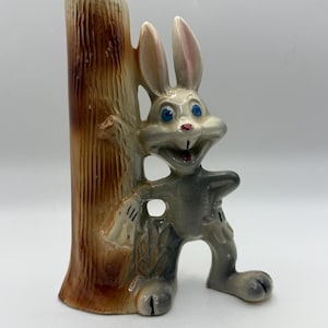 May include: A ceramic figurine of a cartoon rabbit leaning against a tree trunk. The rabbit is gray with blue eyes and pink accents. The tree trunk is brown and tan. The figurine is likely a decorative item or a vase.