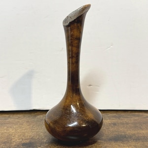 May include: A brown, slender vase with a bulbous base and a flared, angled opening. The vase has a glossy finish and a lighter-colored rim. The vase is displayed on a wooden surface.