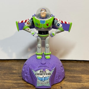May include: A Buzz Lightyear action figure stands on a purple base. The figure is wearing a green, white, and purple spacesuit with a clear helmet. The base has the words "Buzz Lightyear" and "Universe Protection Unit".