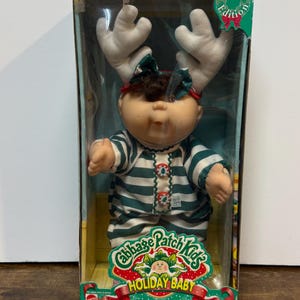 May include: A Cabbage Patch Kids Holiday Baby doll in its original box. The doll wears a green and white striped outfit with red and green buttons. It has white antlers and a green bow. The box reads "Holiday Baby" and "Special Edition."
