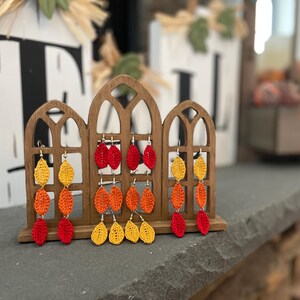 May include: Crocheted leaf earrings in shades of red, orange, and yellow. The earrings are displayed on a wooden stand with a decorative window design.