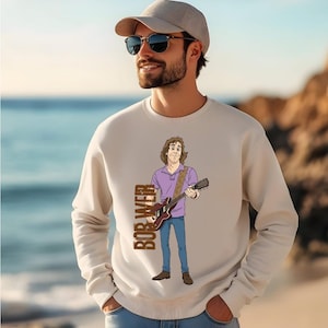 May include: Cream-colored sweatshirt featuring a cartoon illustration of a musician playing a guitar. The illustration includes the text "BOB WEIR" in a vertical brown font. The person in the image is wearing sunglasses and a cap.