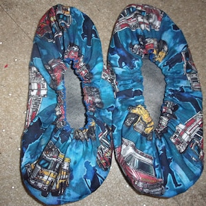 May include: A pair of blue and black fabric slippers with a pattern of cartoon fire trucks and other vehicles. The slippers have a gray lining and a gathered elastic top.