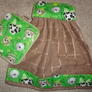 May include: A brown kitchen towel with a green shamrock and dog print border. The towel has a matching green pot holder with a shamrock and dog print. The pot holder has a loop for hanging.