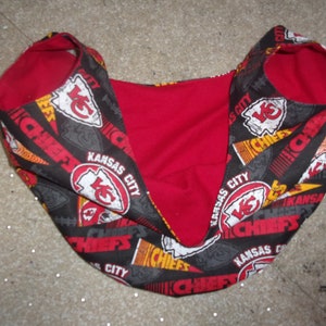K&ouml;nnte beinhalten: Ein roter und schwarzer Stoff-Babysling mit einem Kansas City Chiefs-Muster. Der Sling ist aus weichem, bequemem Stoff gefertigt und perfekt zum Tragen eines Babys.