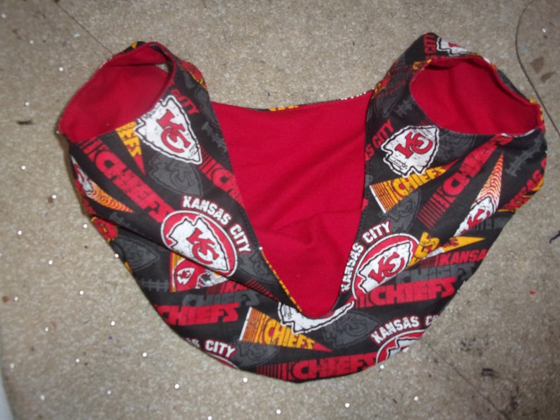 K&ouml;nnte beinhalten: Ein roter und schwarzer Stoff-Babysling mit einem Kansas City Chiefs-Muster. Der Sling ist aus weichem, bequemem Stoff gefertigt und perfekt zum Tragen eines Babys.
