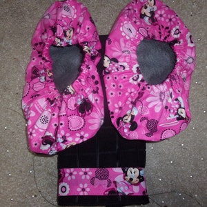 May include: A pair of pink fabric shoe covers with a Minnie Mouse print. The shoe covers are attached to a black towel with a pink Minnie Mouse trim.
