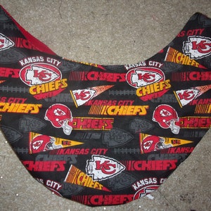 K&ouml;nnte beinhalten: Schwarzer Stoff mit einem sich wiederholenden Muster des Logos der Kansas City Chiefs, den Worten "Kansas City Chiefs" und einem Footballhelm. Das Logo ist rot, gelb und wei&szlig;. Die Worte sind gelb und wei&szlig;. Der Helm ist rot und wei&szlig;.