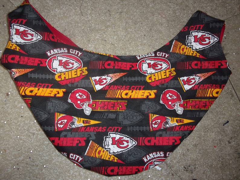 K&ouml;nnte beinhalten: Schwarzer Stoff mit einem sich wiederholenden Muster des Logos der Kansas City Chiefs, den Worten "Kansas City Chiefs" und einem Footballhelm. Das Logo ist rot, gelb und wei&szlig;. Die Worte sind gelb und wei&szlig;. Der Helm ist rot und wei&szlig;.