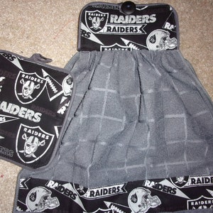 May include: A gray and black kitchen towel set with the Oakland Raiders logo. The towel has a checkered pattern and the pot holder has a black background with white Raiders logos.