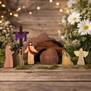 Può includere: Scena pasquale in legno con figure di Gesù, angeli e donne, insieme a una croce e un sepolcro. Sul sepolcro è incisa la scritta "He is Risen!". Le figure sono su una superficie di legno con verde e fiori.