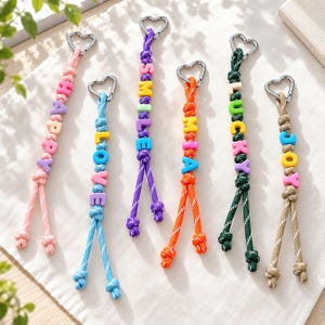 May include: Six colourful paracord keychains with silver heart-shaped carabiners. Each keychain features knotted cords and plastic letter beads spelling out the words HAPPY, LOVE, SMILE, MIA, LUCKY, and JOY in various bright colours and patterns.