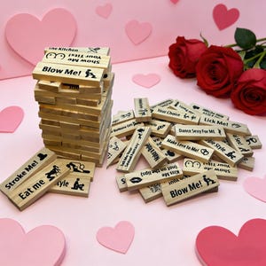 Peut inclure: Un jeu de blocs en bois avec des phrases et des illustrations telles que "Stroke Me!" et "Blow Me!". Les blocs sont dispersés sur une surface rose avec des décorations en forme de cœur et des roses rouges.