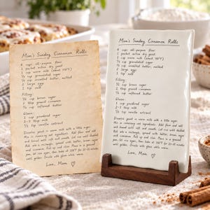 May include: A rectangular white ceramic plaque featuring the printed recipe for Mom's Sunday Cinnamon Rolls, displayed on a dark brown wooden stand next to a matching aged paper print of the same recipe, both listing ingredients and instructions.