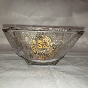 Puede incluir: Un cuenco de vidrio transparente con un diseño dorado de un pájaro con una cola en forma de abanico y una estrella encima. El cuenco tiene una base cuadrada y una parte superior redondeada.
