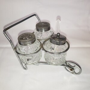 May include: A silver metal condiment holder shaped like a wheelbarrow with three glass jars with silver lids. The jars have a textured, diamond-shaped pattern.