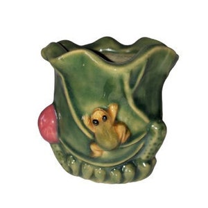 May include: A green ceramic planter shaped like a leaf with a small frog figurine on it. The planter has a pink accent on the side.