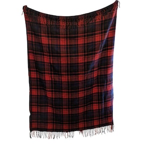 May include: A rectangular red, black, and blue plaid blanket with fringe along the bottom edge. The blanket is hanging against a white background. The plaid pattern is a classic design, suitable for a variety of uses.