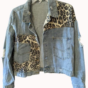 May include: A light blue denim jacket featuring distressed detailing and contrasting leopard print panels on the upper back, shoulders, and front pockets. The jacket includes a classic collar, long sleeves, and a button-down front closure.