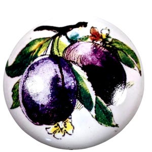 May include: A round white ceramic knob featuring a detailed illustration of two dark purple plums with green leaves and a small yellow flower. The surface is smooth and glossy, showcasing a botanical print centered on the circular face of the hardware.