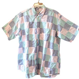 May include: A short-sleeved button-down shirt featuring a patchwork design of seersucker fabric squares in light pink, mint green, and navy blue stripes. The shirt has a standard collar, a front button placket, and a single chest pocket.