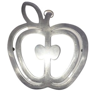 May include: A silver-colored metal apple-shaped trivet with a leaf and a small ring for hanging. The trivet has a cutout design, with a heart shape in the center of the apple. The metal has a brushed finish.