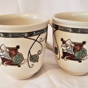 May include: Two white ceramic mugs with a floral design. The mugs have a brown, green, and blue floral pattern with gold accents.