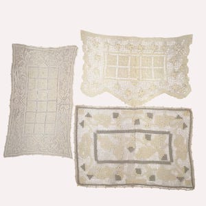 May include: Three rectangular off-white crochet doilies featuring intricate needlework patterns. One doily has a central grid, another has a scalloped edge with floral motifs, and the third displays a geometric border with leaf-like designs.