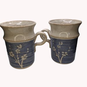 May include: Two ceramic mugs with a two-tone design. The top portion is a light beige, while the bottom is a textured blue with a white floral pattern. Each mug has a handle and a flared rim, perfect for enjoying your favorite beverage.