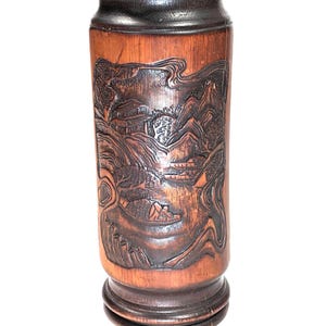 May include: A cylindrical dark brown wooden brush pot featuring intricate relief carvings of a landscape scene with mountains, trees, a small building, and a person in a boat on water. The rim has a geometric triangular pattern.