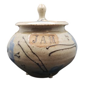 May include: A ceramic jam jar with a lid. The jar is beige with blue and brown accents. The word "JAM" is embossed on the front. The jar has a rounded shape and three small feet.