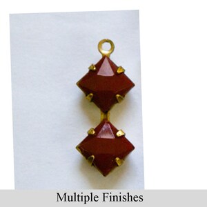 May include: A vertical pendant featuring two square-cut, deep red stones set in gold-toned metal prongs. The stones are arranged in a stack, with a small circular loop at the top for attachment. The text "Multiple Finishes" appears below.