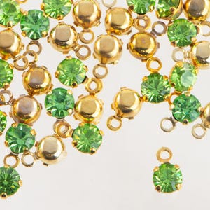 May include: Close-up of scattered gold-colored metal settings with round, faceted, light green gemstones. The settings have small loops for attachment. The gemstones are a vibrant, bright green color. The background is white.